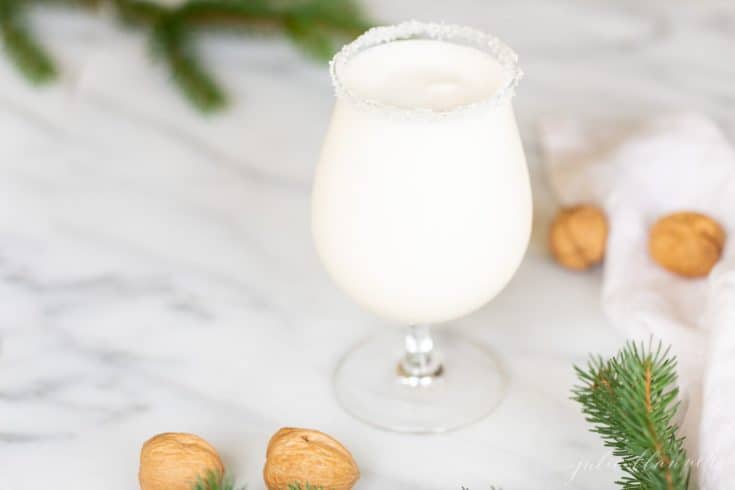 一个透明的高脚杯，盛满白色的圣诞鸡尾酒，顶部有糖边、榛子和绿叶。