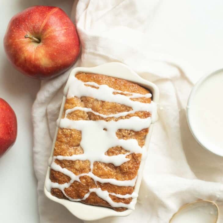 一个装着苹果面包的小面包盘，上面撒着白色糖衣，四周放着苹果和亚麻餐巾。