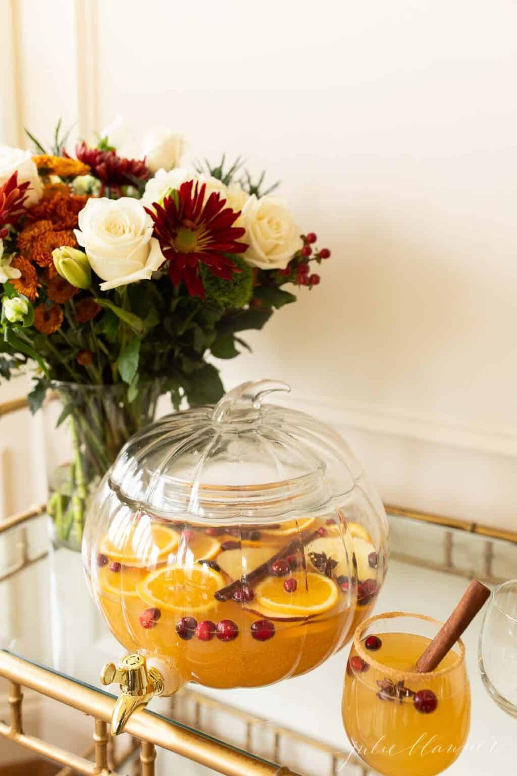 一个透明的玻璃南瓜饮料分配器装满了苹果酒桑格利亚，背景是鲜花。