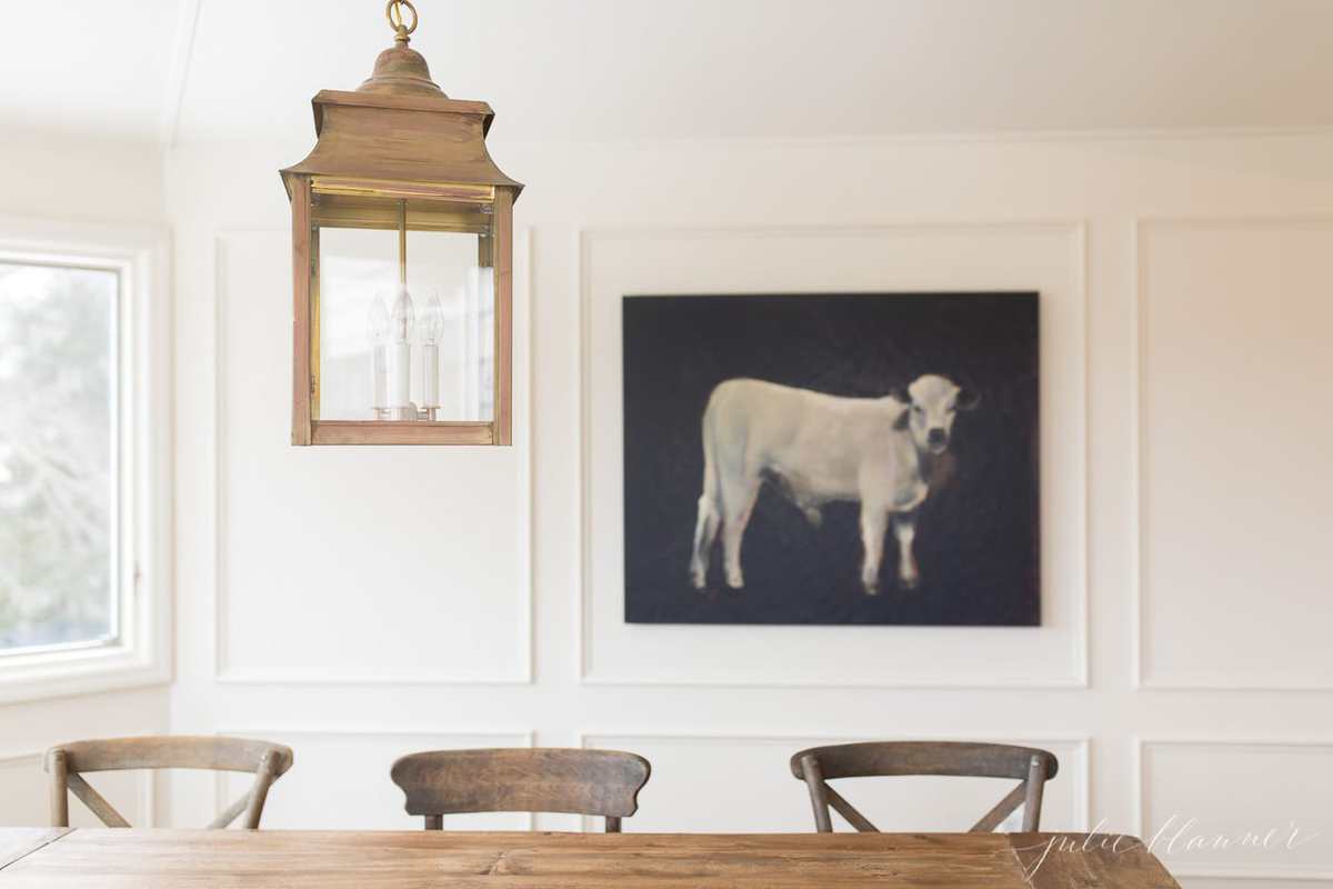 厨房旁边是一间白色的早餐室，有一张农场桌子，黄铜灯笼，背景是一幅奶牛的画。