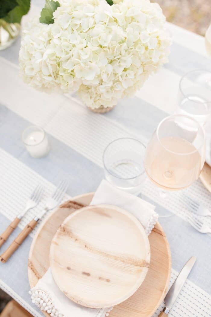 在蓝白相间的桌布上摆放一个木制的盘子和餐具，上面放着白色的花，供露天晚餐用。