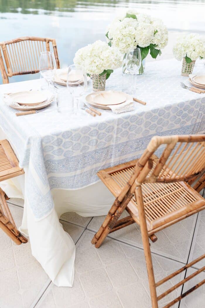 一个露天的餐桌，白色的花朵和蓝白相间的桌布，水为背景。