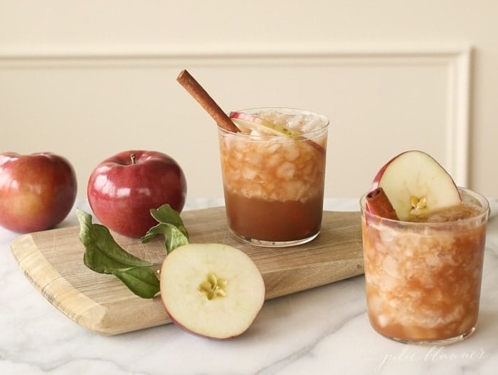 一杯苹果感恩节鸡尾酒，装在透明玻璃杯里，配肉桂棒和苹果片，旁边放满红苹果。