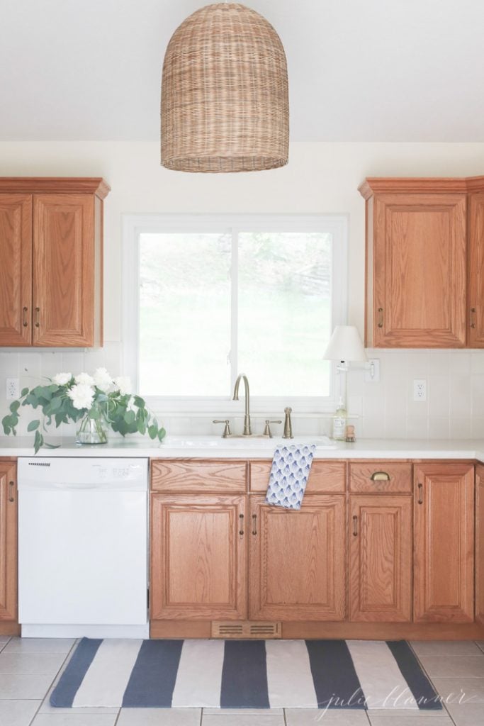 90年代的厨房与橡木橱柜看起来现代和清新藤灯具，香槟青铜水龙头，和蓝白色条纹地毯在水槽前。
