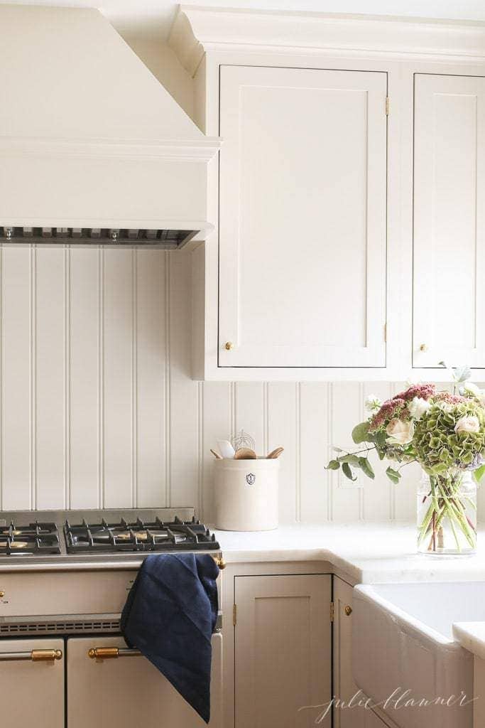 在奶油色的厨房里，水槽前面系着围裙，旁边放着一瓶花。