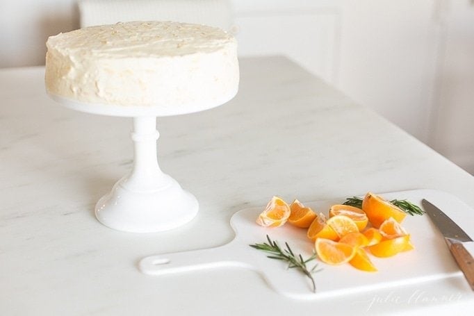 白色底座上的桔子蛋糕，旁边的砧板上放着橘子片和迷迭香。gydF4y2Ba