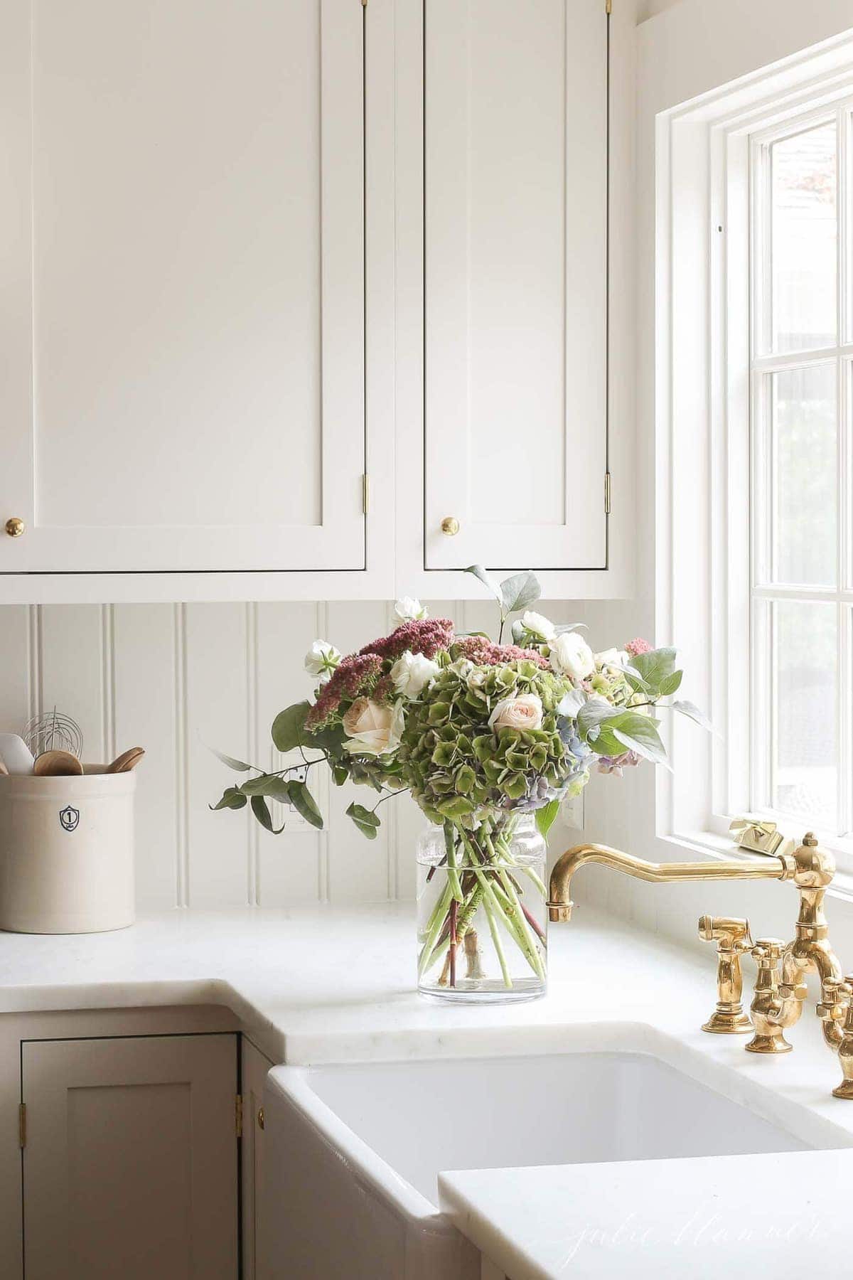 一个明亮而舒适的家庭厨房，带有白色橱柜，黄铜龙头和水槽旁的花瓶。