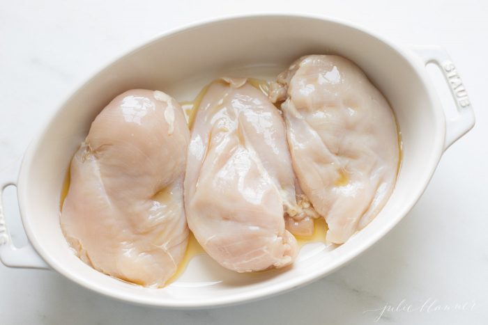 一个白色椭圆形的菜，上面有三个未煮过的鸡胸肉，橄榄油。GydF4y2Ba