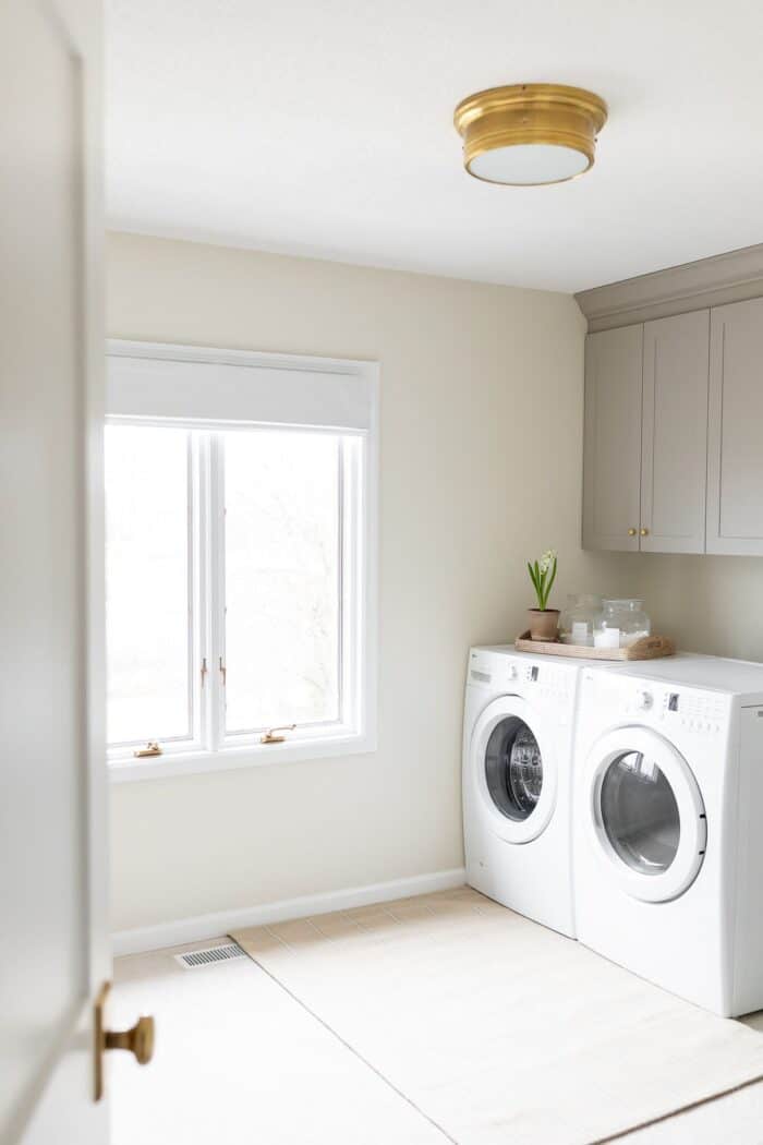 一间大的洗衣房，有一扇窗户，灰色的柜子放在洗衣机和烘干机前面。