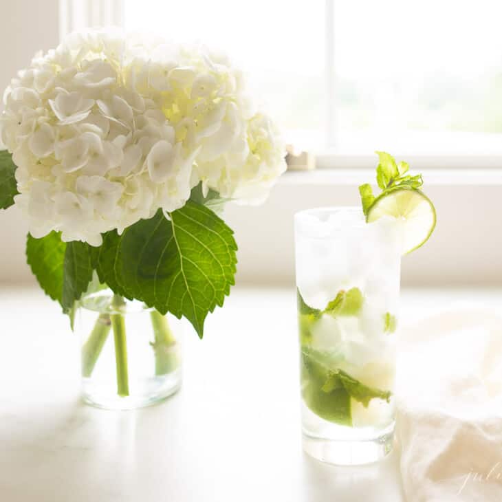 白色的工作台面上有一个插着绣球花的花瓶和一个高的透明玻璃和莫吉托。gydF4y2Ba