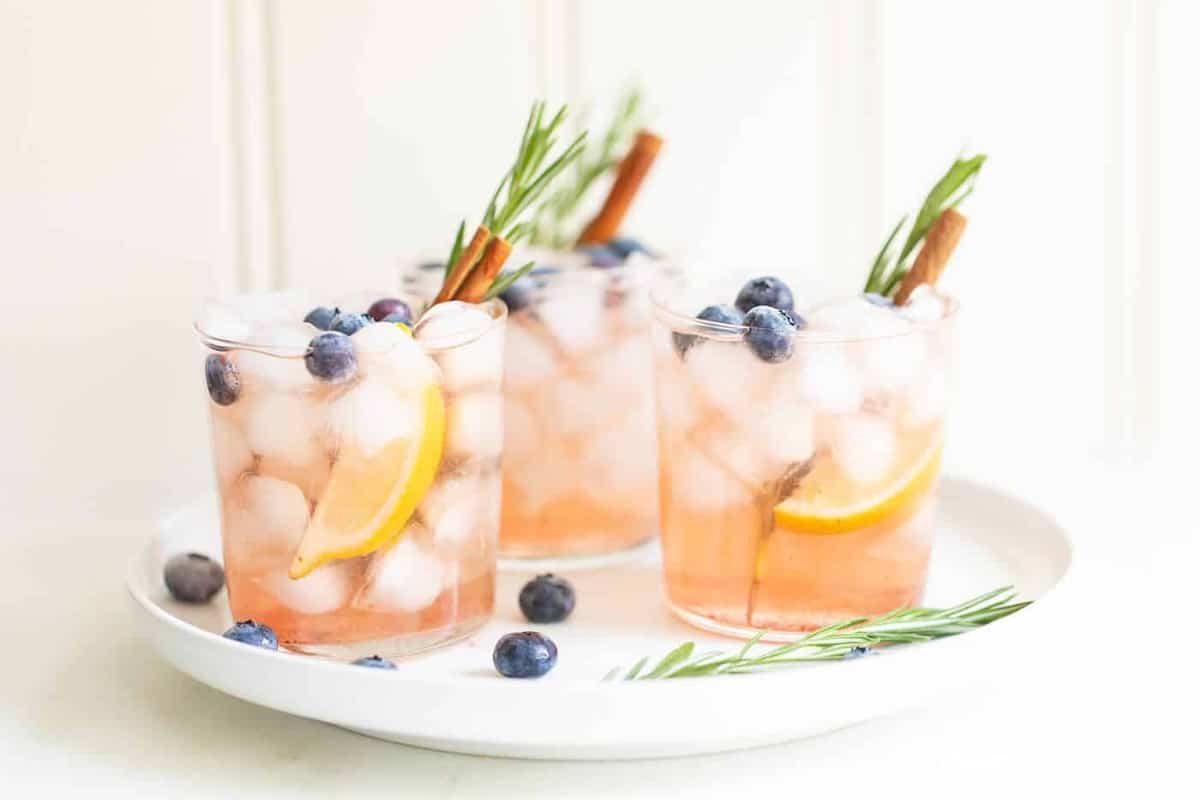 三击鸡尾酒配上迷迭香、蓝莓、橙子和肉桂棒，放在一个白色的盘子里。