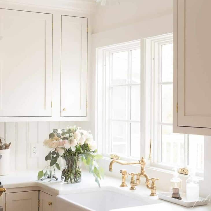 一个白色的厨房，有农舍水槽，黄铜厨房龙头和一瓶鲜花到侧面。