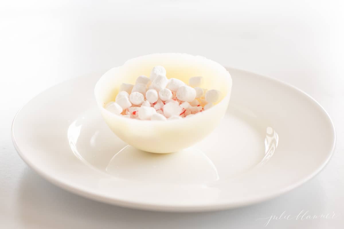 一个白巧克力热可可炸弹在一个白色的盘子，一分为二显示棉花糖和混合。gydF4y2Ba