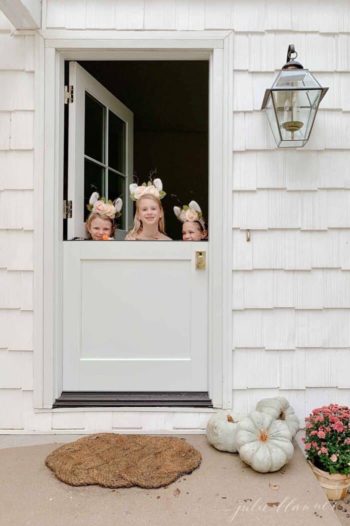 白色小屋的外面有一扇白色的荷兰门，门把手是黄铜的，三个小女孩从门的下半部分探出头来。