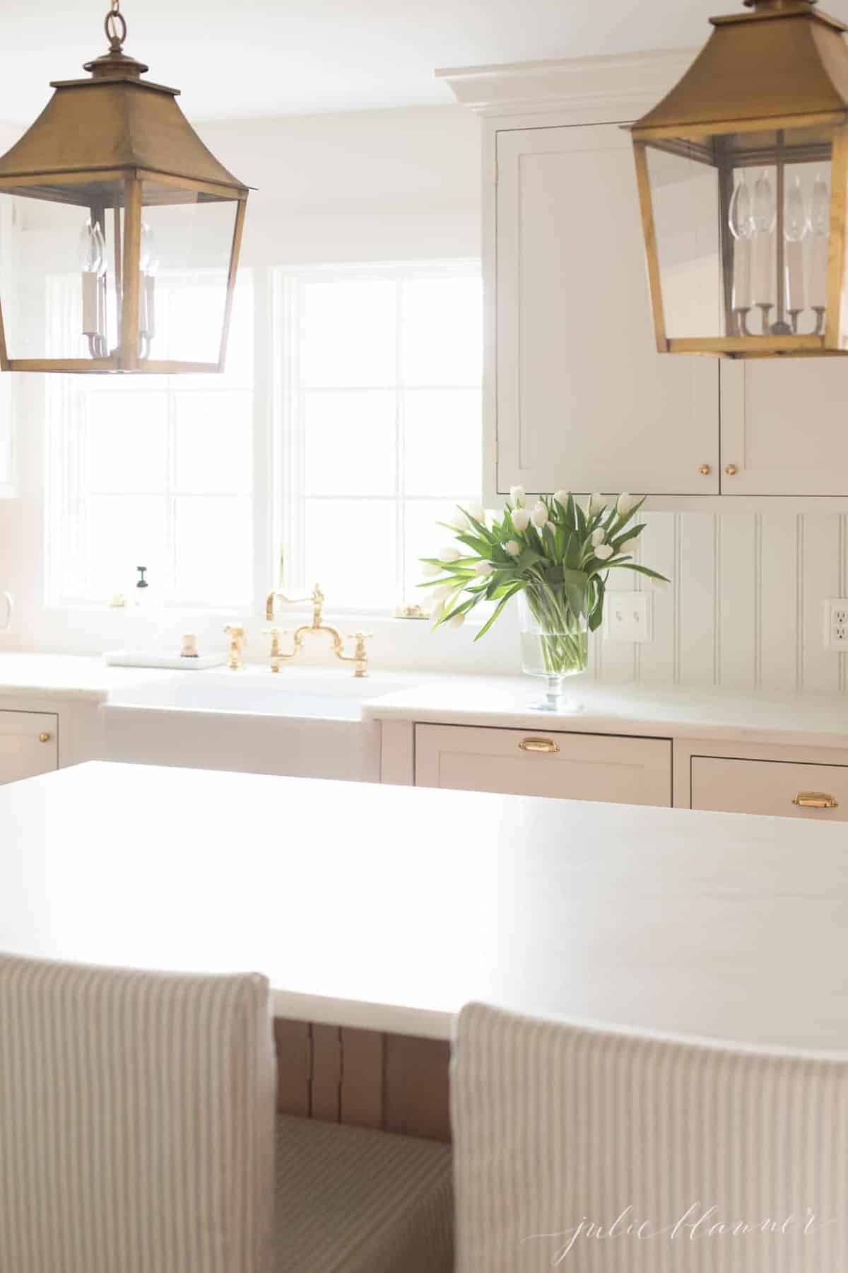 白色的厨房配有摇床风格的橱柜，黄铜灯笼悬挂在岛上。