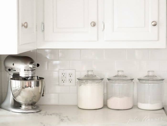 白色的厨房里有大理石台面，一个立式搅拌机和装满烘焙用品的玻璃罐。