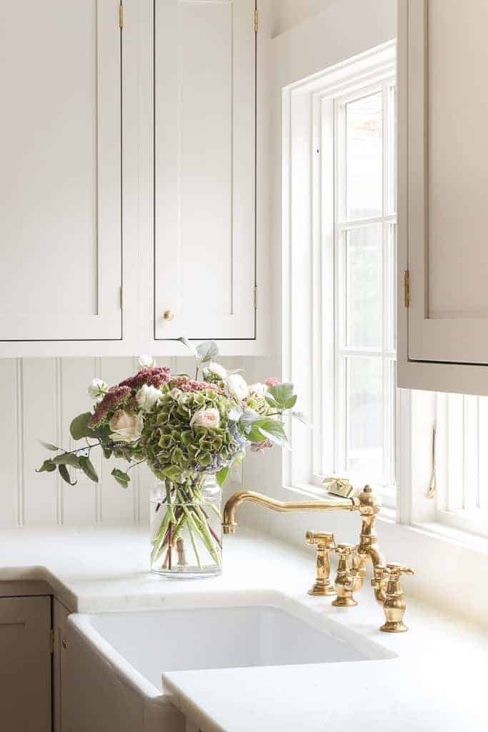 白色的厨房里有摇瓶柜和大理石柜台，水池边放着一瓶花。