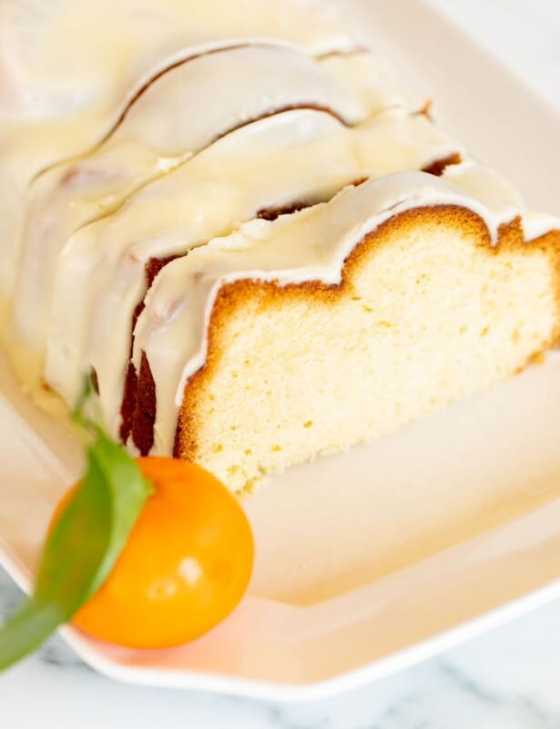 在结霜的一个切的橙色磅蛋糕在白色盛肉盘，桔子在背景中。