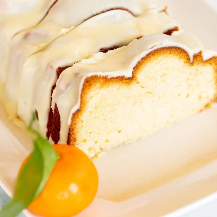白色的浅盘上放着一层裹着糖霜的切成薄片的橘色磅蛋糕，背景是橘子。gydF4y2Ba