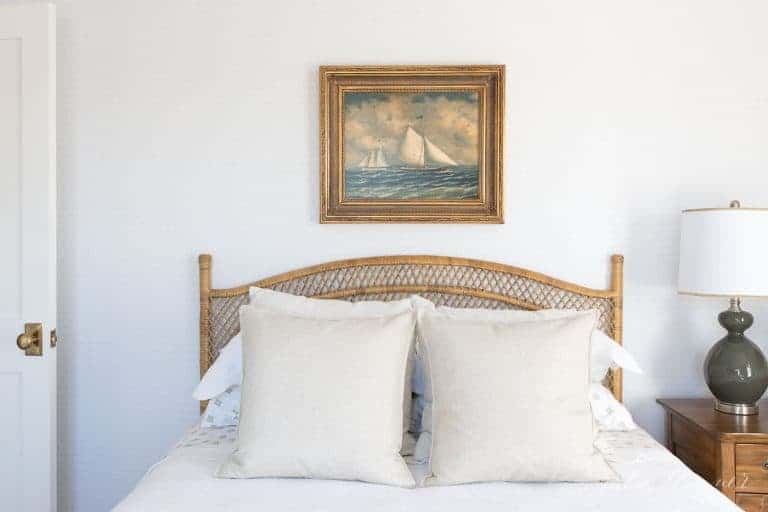 一间带有中性床上用品的卧室，一张藤床，床头板上方是帆船艺术品。