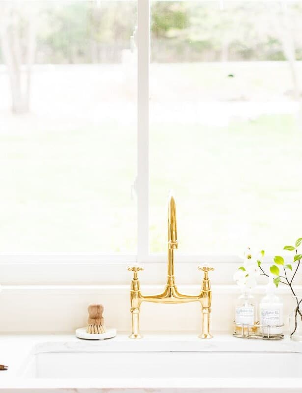 一个白色的厨房水槽区域，带有黄铜桥龙头，水槽上的窗户和花瓶到侧面。