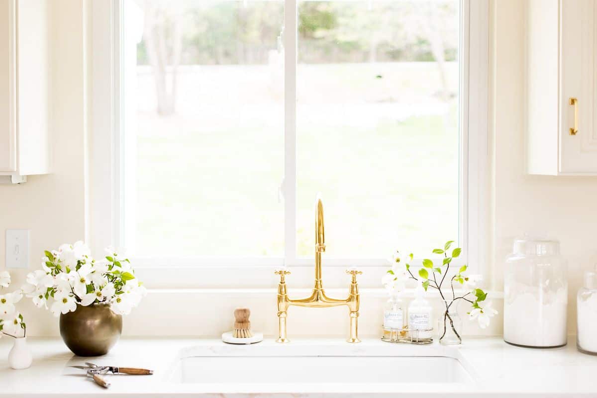厨房里白色的水槽区有一个黄铜桥式水龙头，水槽上方有一扇窗，旁边放着一瓶花。