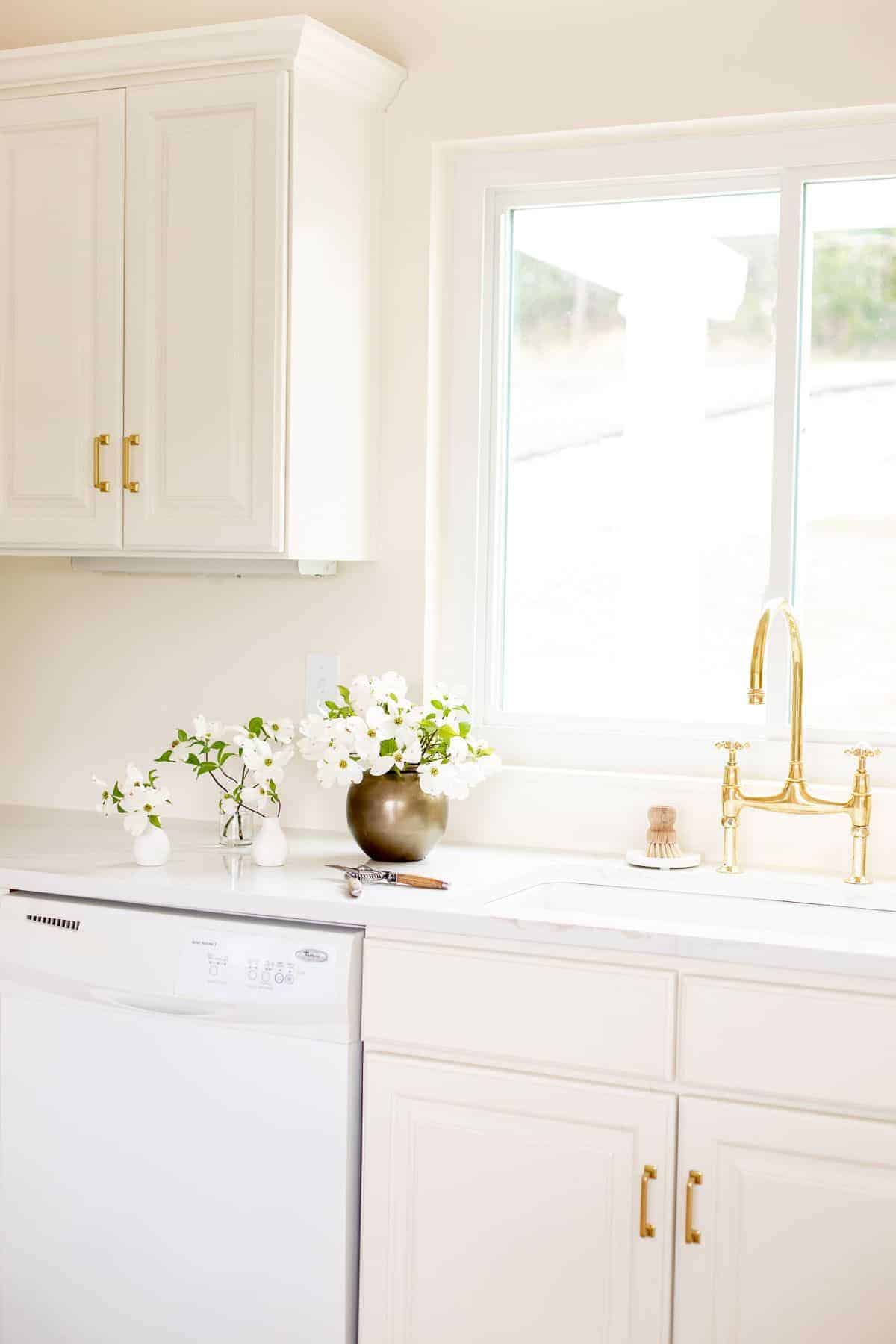 厨房里白色的水槽区有一个黄铜桥式水龙头，水槽上方有一扇窗，旁边放着一瓶花。