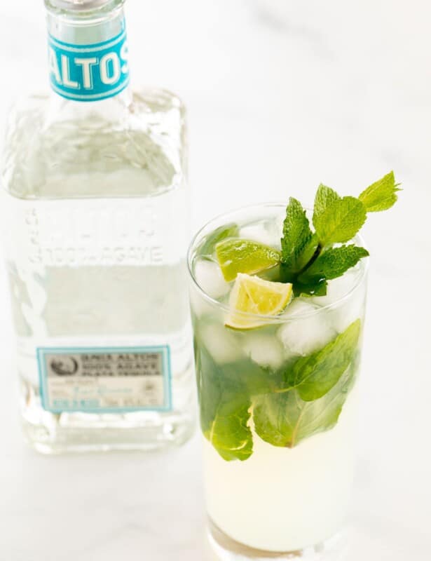 在白色表面上的透明玻璃杯中的龙舌兰酒莫吉托（Tequila Mojito），上面饰有柠檬楔子和新鲜薄荷，背景中的龙舌兰酒。