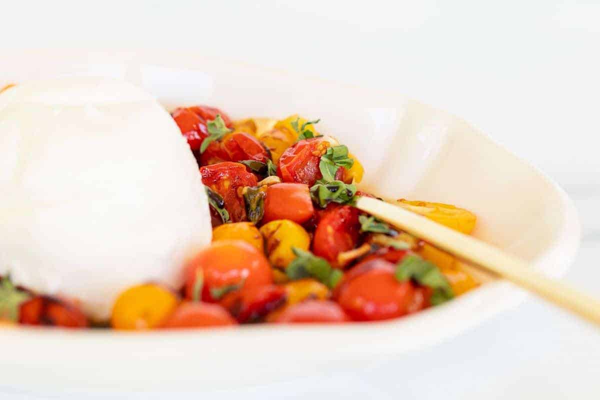 一块烤布拉塔奶酪放在一个白色的碗里，里面放着烤樱桃番茄，旁边放着金汤匙。gydF4y2Ba