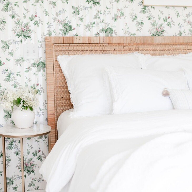 一间带花卉墙纸的卧室，藤质床头板，白色的床上用品和金色和大理石边桌。