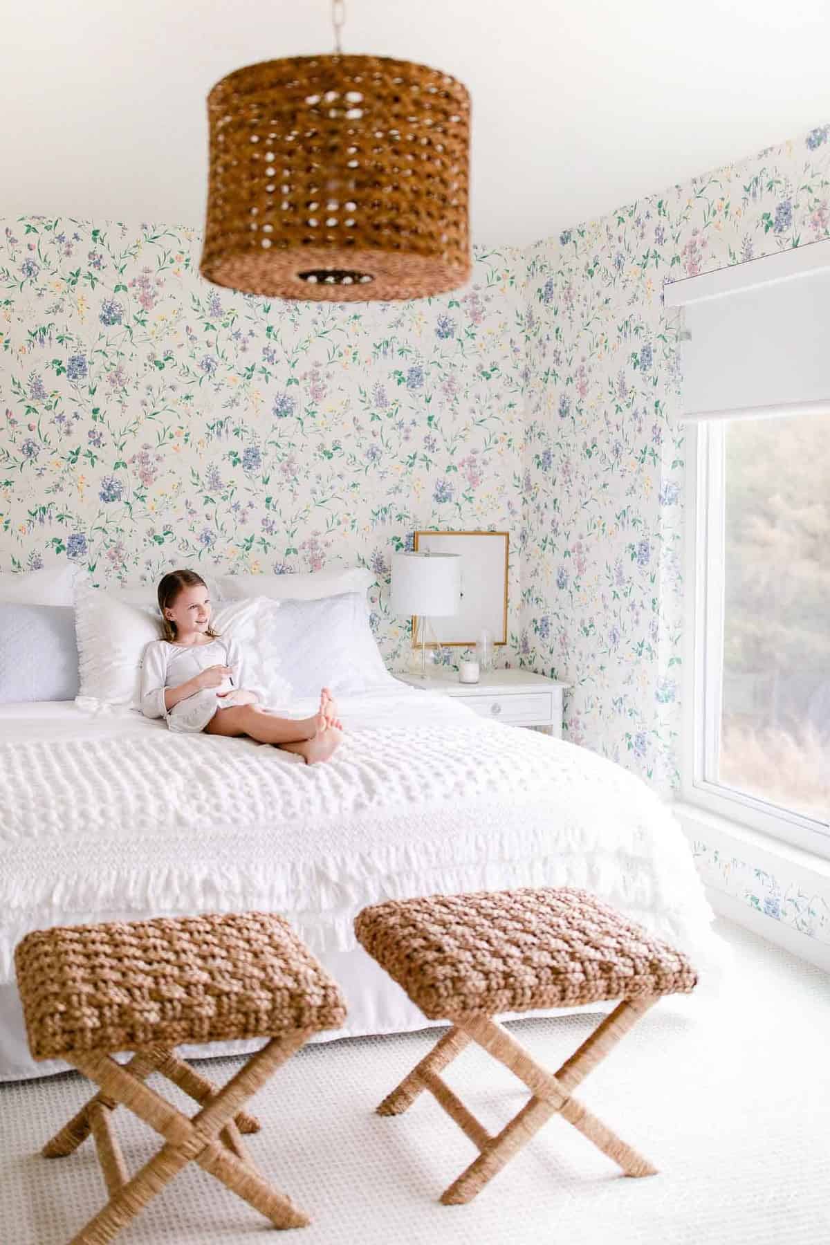 一间带花卉墙纸装饰的白色卧室，一个小女孩坐在床上。