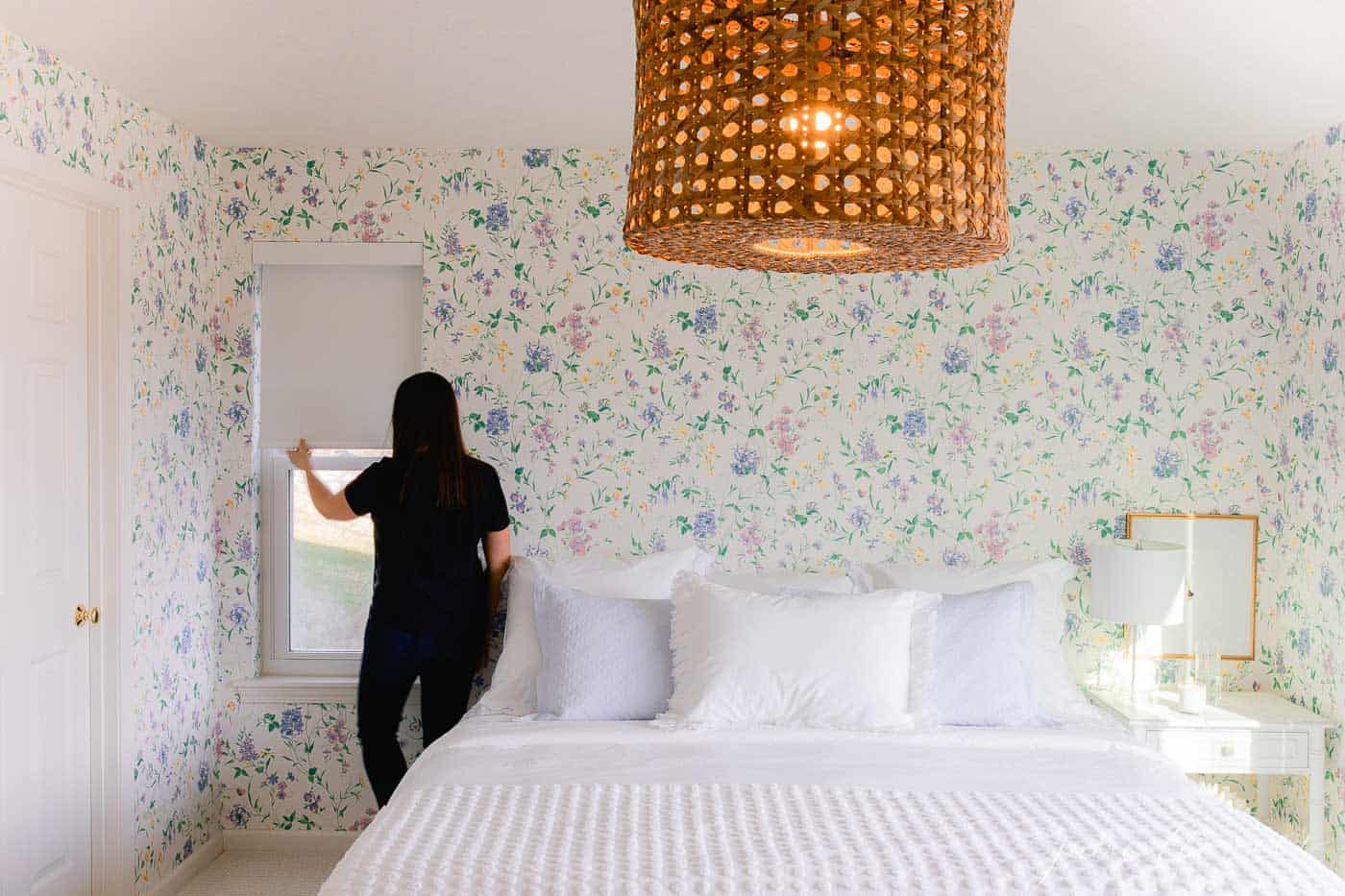 一间带有木制装饰的白色卧室和一个陈述花卉墙纸，一个女人将阴影拉到侧面。